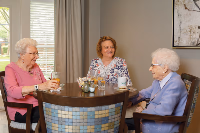 Three elderly women sitting around a round wooden table in a well-lit room, enjoying drinks and snacks while engaging in conversation. The room has a window with blinds and curtains, and a framed artwork on the wall.