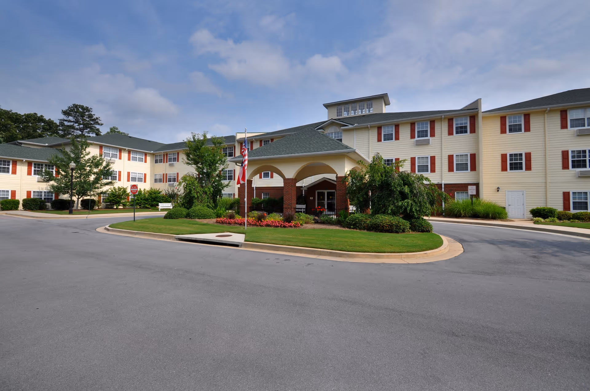 Exterior view of Creekside Pines Retirement Community showing a large three-story building with beige siding and red shutters. The entrance features a covered driveway with brick pillars, surrounded by landscaped greenery and flowers. An American flag is displayed near the entrance under a partly cloudy sky.