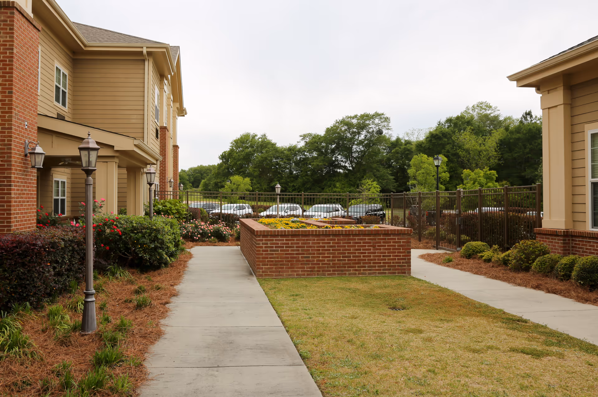 Outdoor view of a senior living facility showing a paved walkway between two buildings with brick and beige siding. There are lamp posts, landscaped bushes, flowers, and a raised brick flower bed in the center. Trees and parked cars are visible in the background under a cloudy sky.