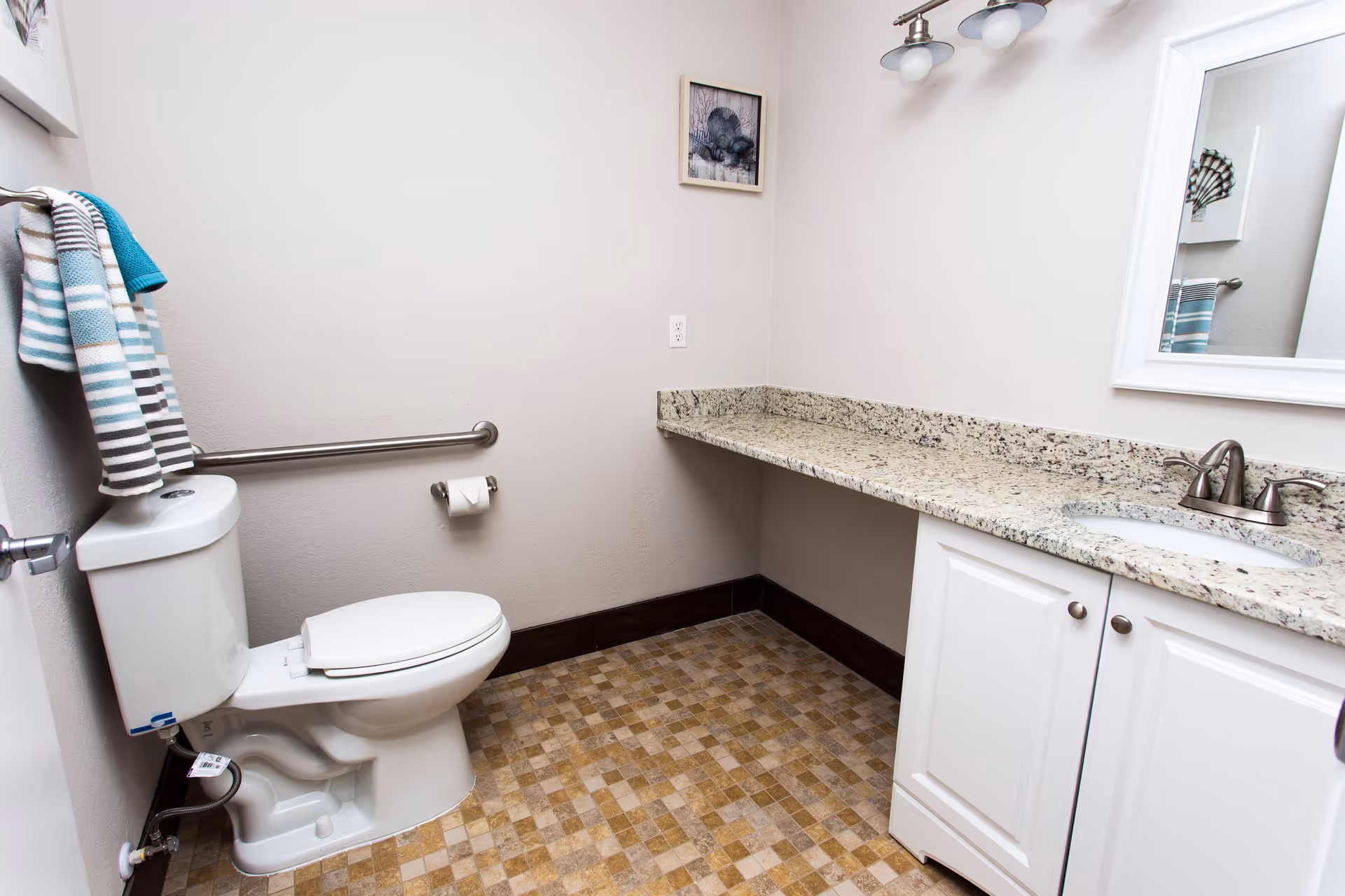 A clean and accessible bathroom with a white toilet, a granite countertop with an under-mount sink, a large mirror, and a towel rack holding striped towels. The floor has a checkered tile pattern in shades of brown and beige. There is a safety grab bar next to the toilet and a small framed picture on the wall.