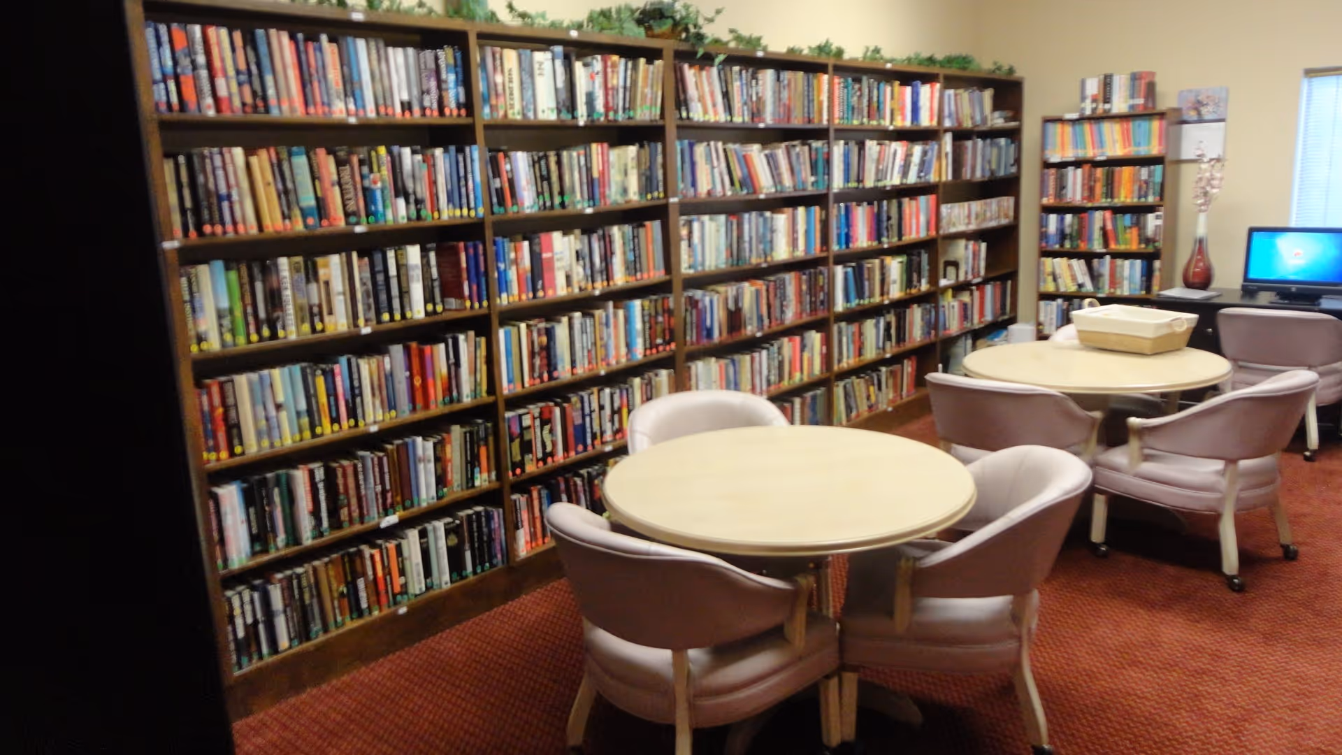 Community library room with wall-to-wall bookshelves, round tables and upholstered chairs.