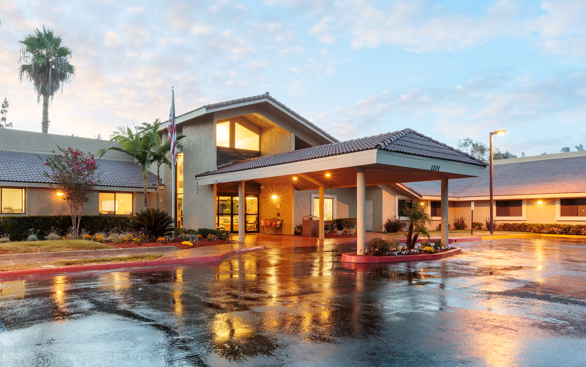 Exterior view of San Diego Post-Acute Center (SDPAC) building at dusk with wet pavement reflecting lights, a covered entrance supported by columns, palm trees, and landscaped flower beds.