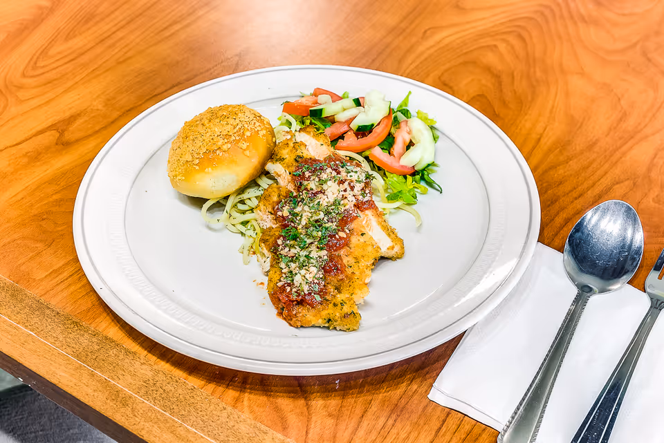 A plate with breaded chicken topped with sauce and herbs, pasta, a salad and a dinner roll on a wooden table next to utensils.