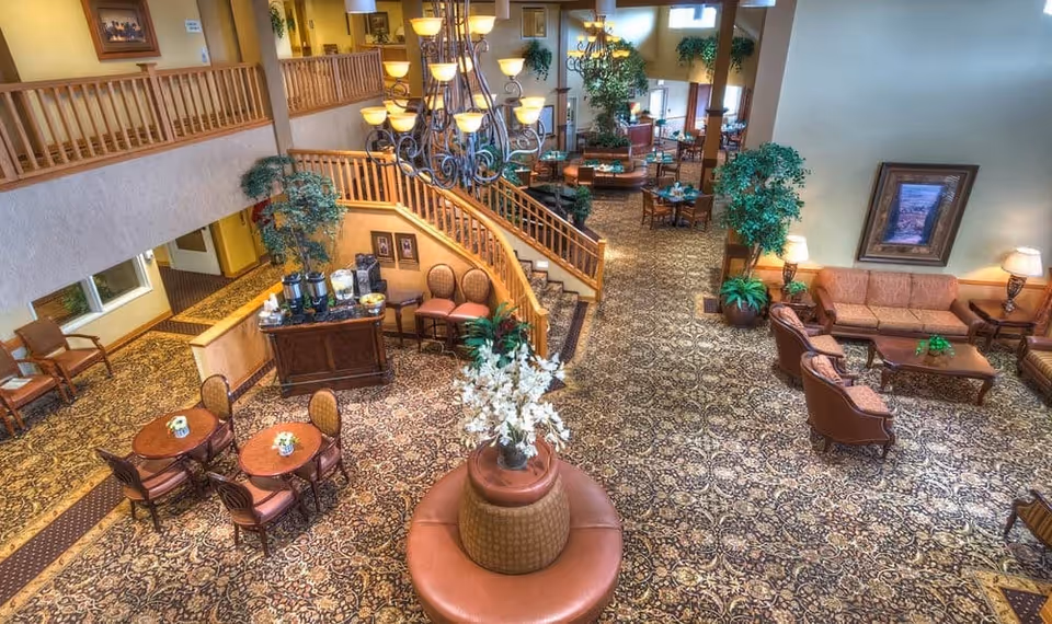 Spacious, warmly decorated senior living common area with seating groups, small dining tables, a staircase, chandelier, and plants.