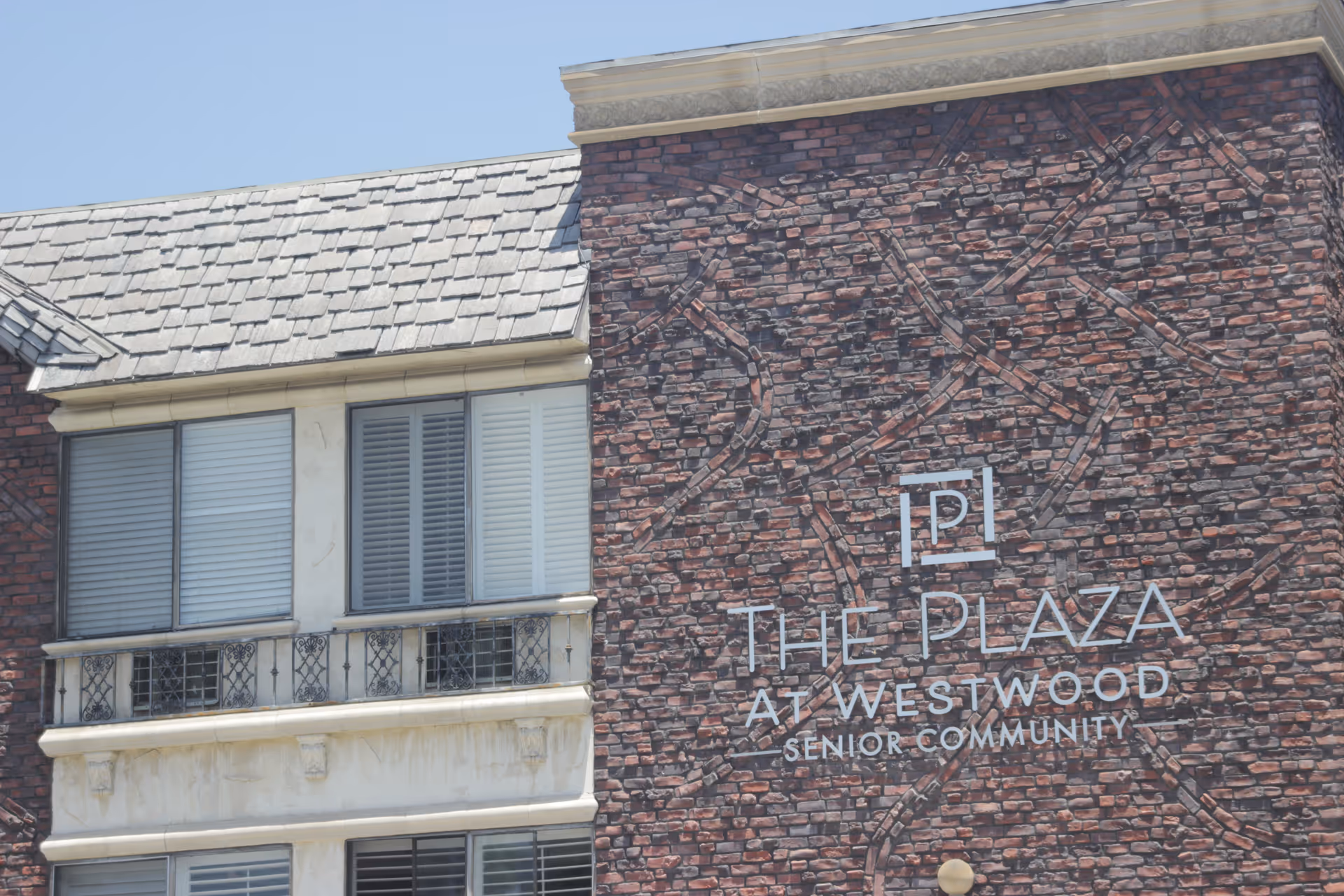 Exterior view of a senior living facility building with a brick facade and a sign that reads 'The Plaza At Westwood Senior Community'. The building has windows with shutters and a shingled roof under a clear blue sky.