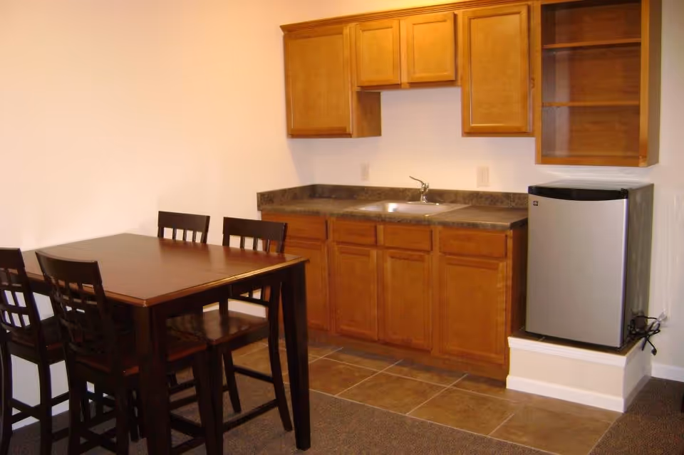 A small kitchen area with wooden cabinets, a countertop with a sink, a mini refrigerator, and a dark wooden dining table with four matching chairs.