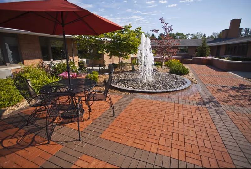 Courtyard with brick pavers, a central fountain, and a metal table with chairs under a red umbrella beside a single-story building.