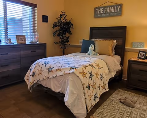 Sunny bedroom with a twin bed covered by starfish-print bedding, wooden headboard, matching dresser and nightstand, a potted plant, and a wall sign reading 'THE FAMILY'.