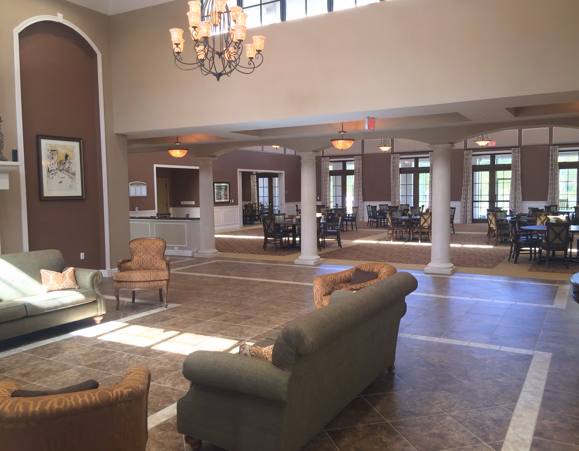 A spacious and well-lit common area in a senior living facility featuring comfortable seating with sofas and armchairs in the foreground, a chandelier hanging from the high ceiling, and a dining area with multiple tables and chairs in the background separated by white columns.