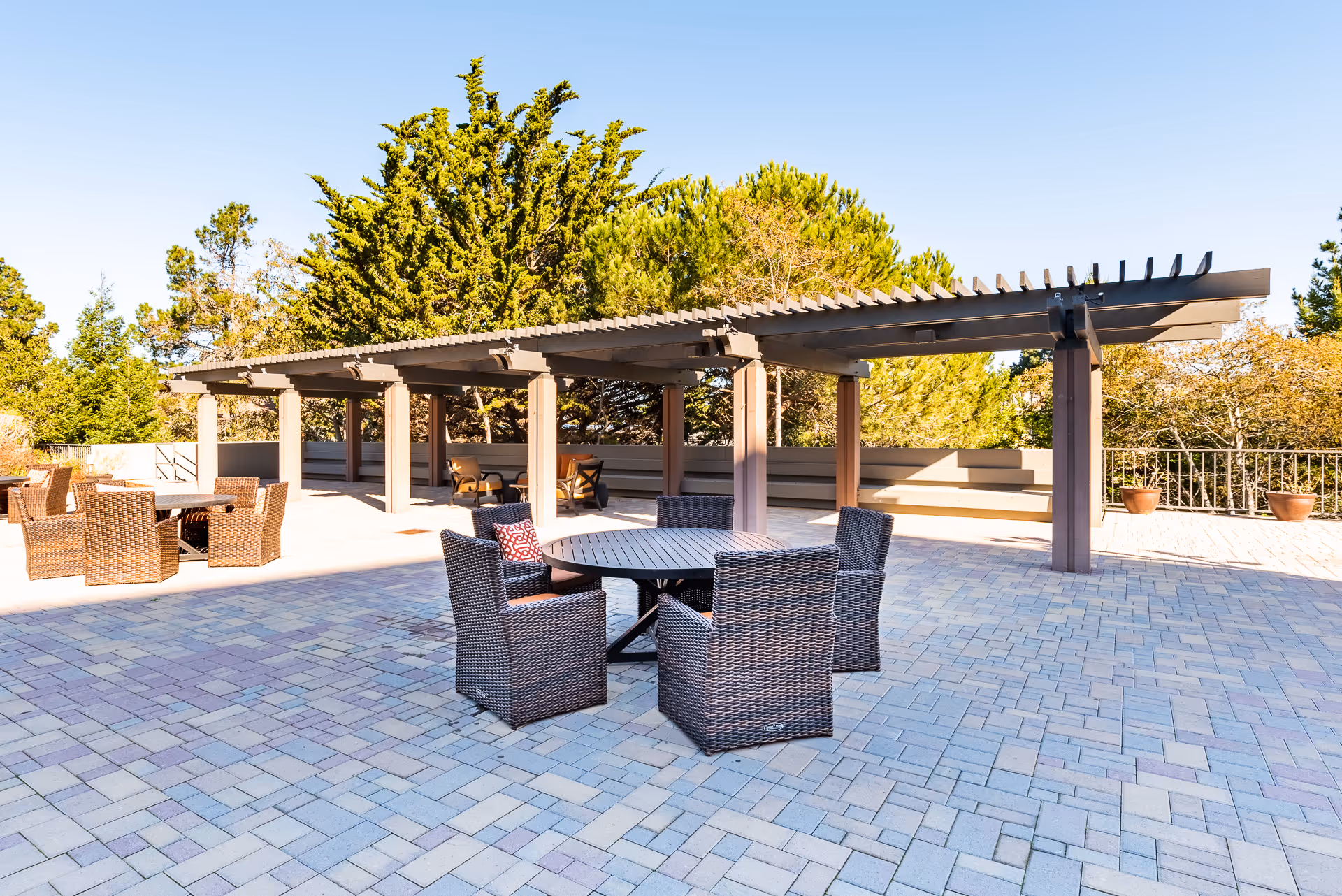 Sunlit outdoor terrace with a pergola, wicker dining chairs and a round table on a paved patio against a backdrop of trees.