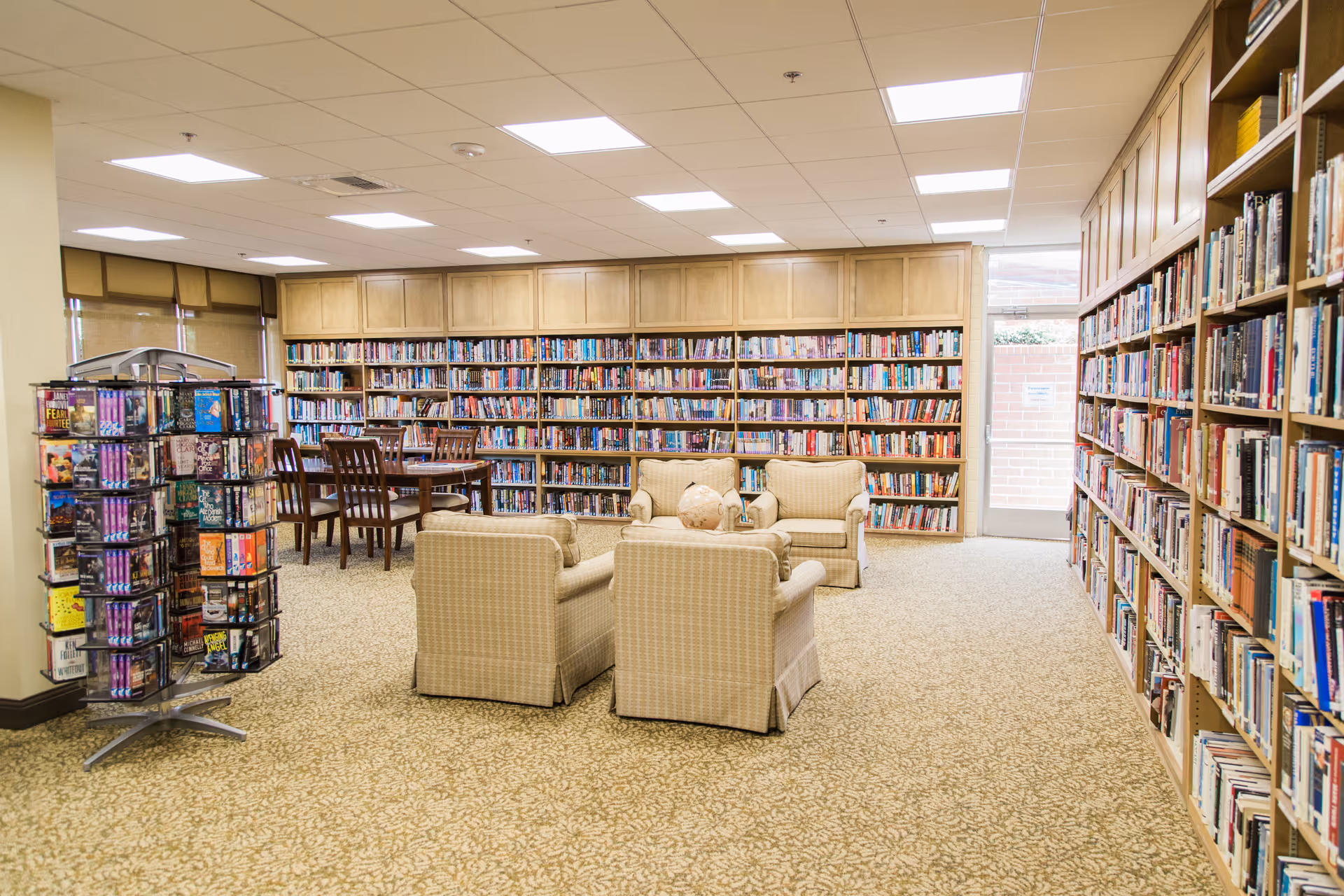 A bright interior library/reading room with bookshelves along the walls, a seating area of upholstered armchairs and a table, and a rotating display rack.