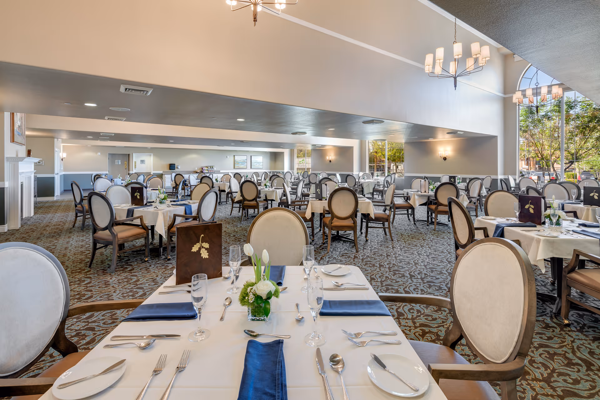A spacious and elegant dining room with multiple round tables covered in white tablecloths and set with silverware, glasses, and navy blue napkins. The chairs have cushioned seats and backs with wooden frames. The room features large windows letting in natural light and decorative light fixtures hanging from the ceiling. The carpet has a patterned design, and there are small floral centerpieces on the tables.