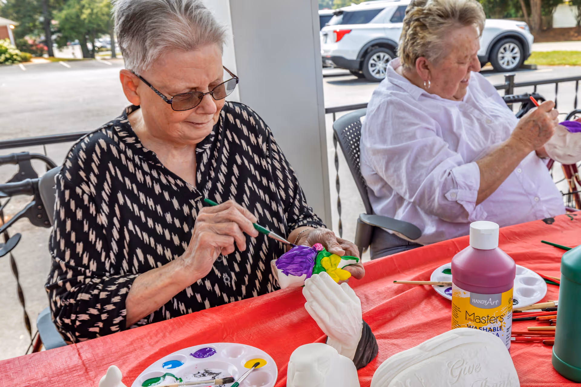Two elderly women sitting at a table outdoors, painting ceramic figurines with colorful paints. One woman is focused on painting a purple and yellow flower, while the other is painting a white ceramic piece. Various paint bottles and brushes are on the red tablecloth-covered table.