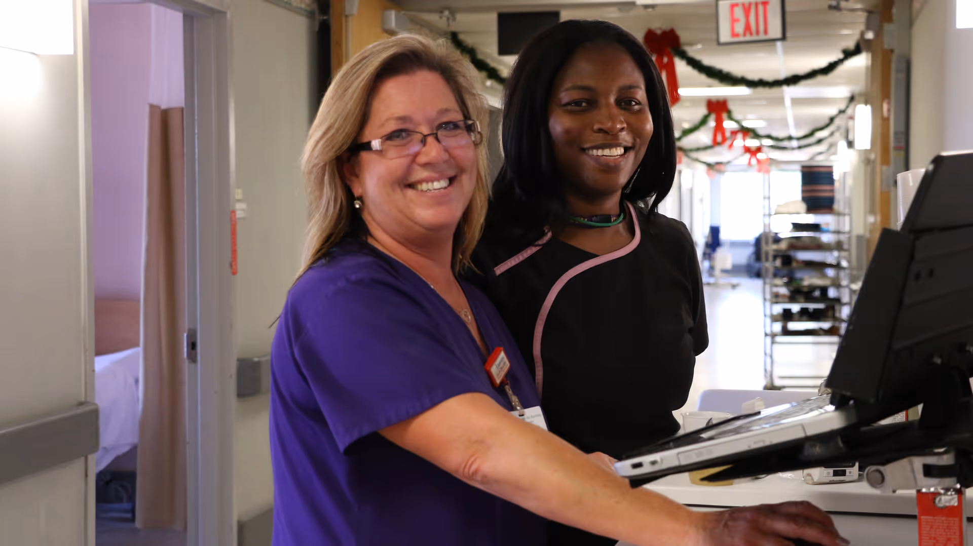 Two healthcare workers smiling and standing together at a computer workstation in a hallway decorated with holiday garlands and red bows.