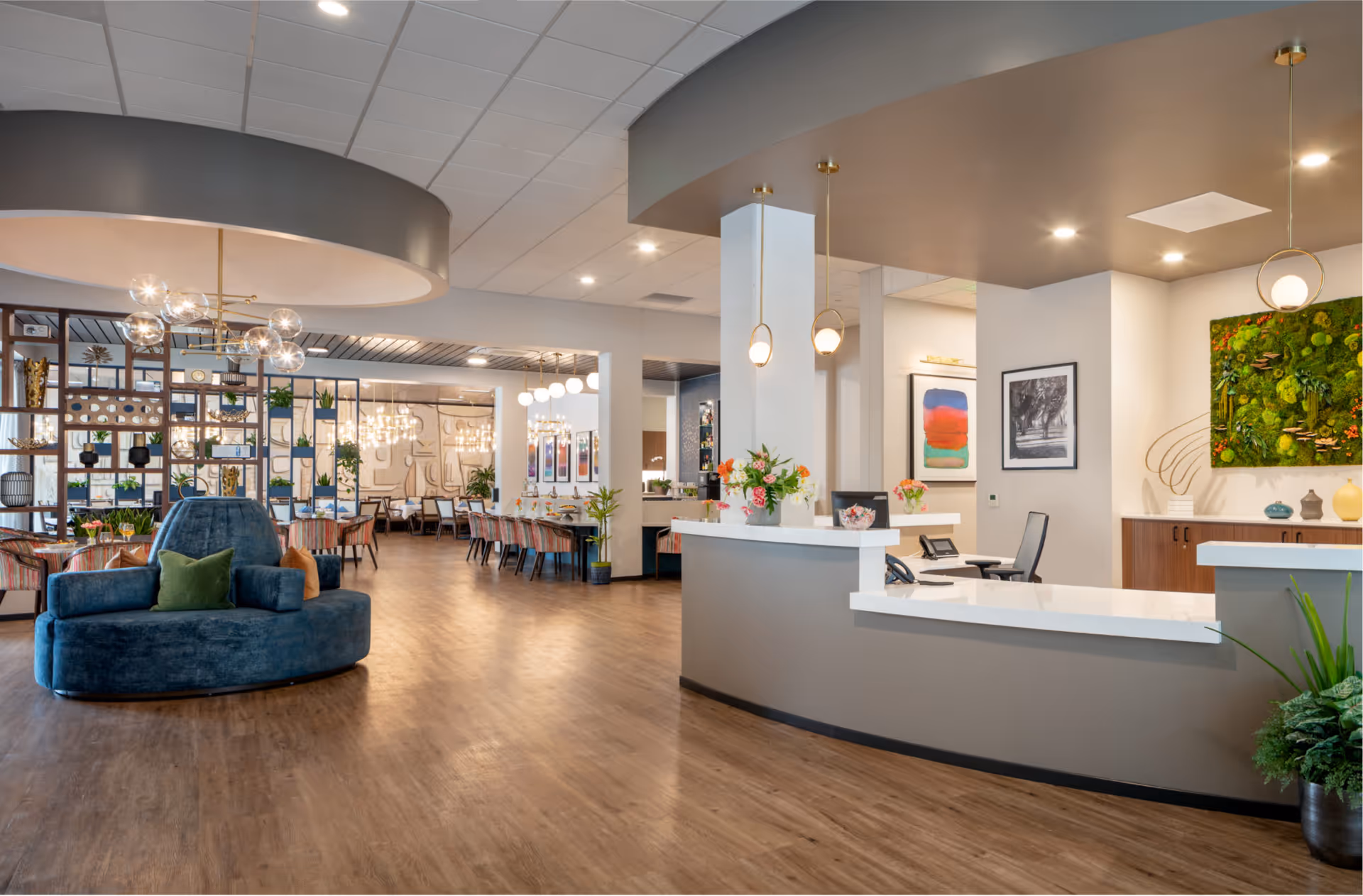 Modern senior living facility interior showing a reception desk, lounge seating, and a dining area with tables and decorative shelving.