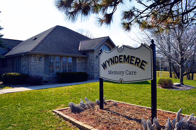 Exterior view of a single-story brick building with a dark shingled roof, surrounded by green grass and trees. In the foreground, there is a sign that reads 'WYNDEMERE Memory Care'.