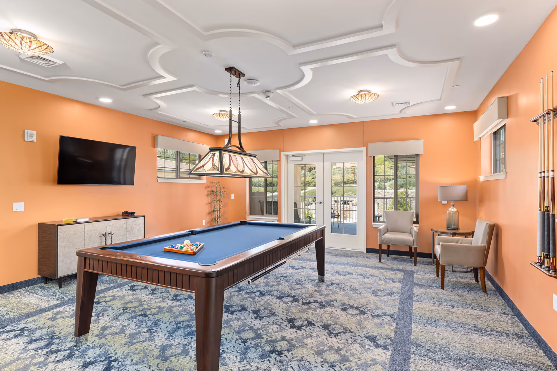 A billiards room featuring a blue pool table, an orange accent wall, a flat-screen TV, and two chairs beside a small table with a lamp. Large windows provide natural light and a view of the outdoors.