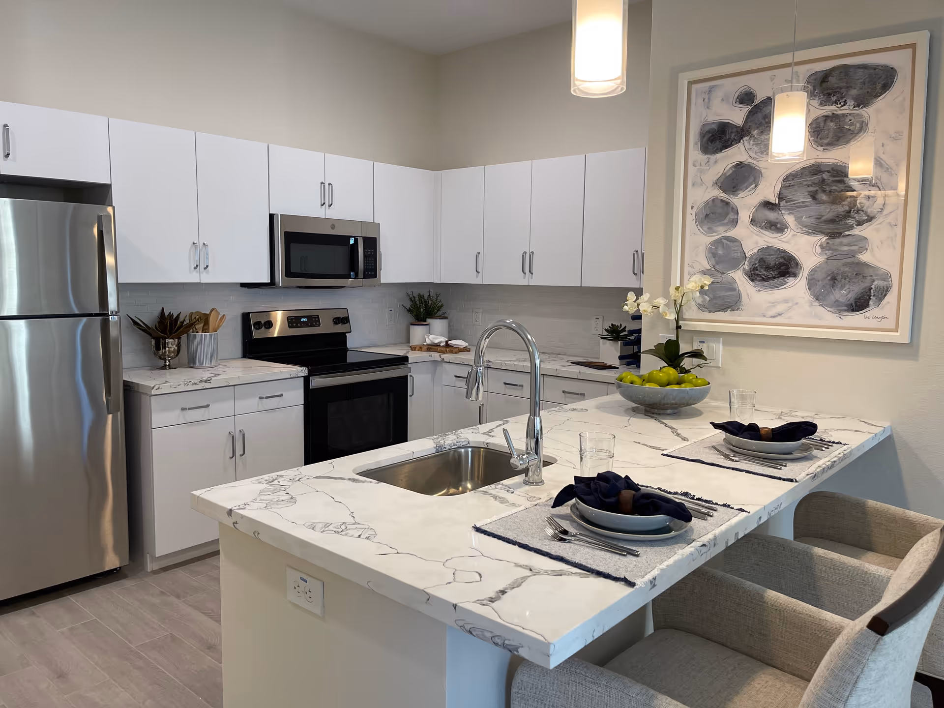 Modern kitchen with white cabinets, stainless steel refrigerator, oven, and microwave. A marble countertop island with a sink and two place settings with plates, napkins, and silverware. A bowl of green apples and a white orchid plant are on the counter. Abstract black and gray artwork hangs on the wall, and two pendant lights hang above the island.