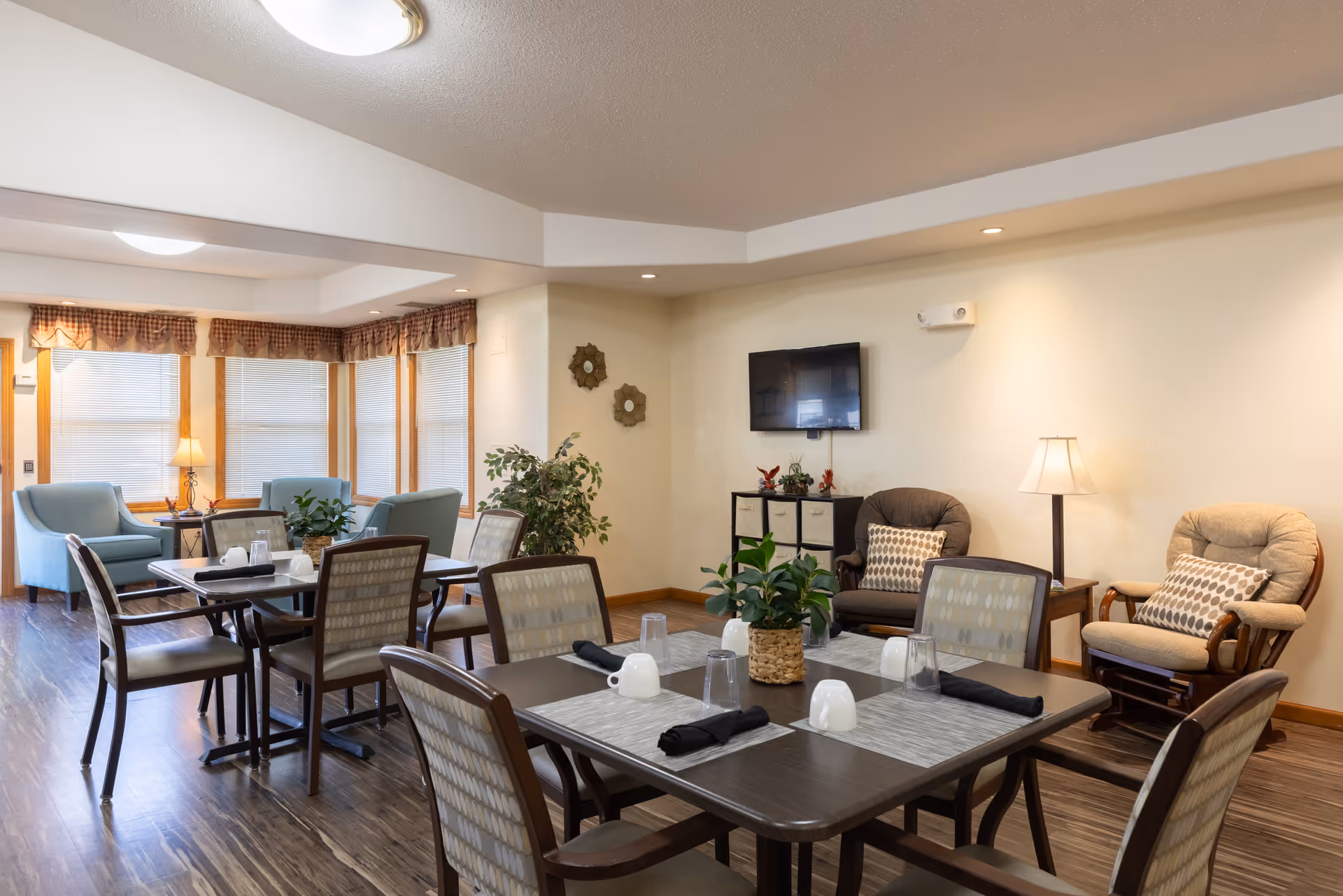 A bright communal dining and sitting area with set dining tables, upholstered chairs, armchairs, plants, and a wall-mounted TV.