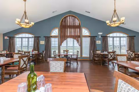 Dining room with multiple wooden tables and chairs arranged neatly. Large arched windows with curtains allow natural light to fill the room. Two chandeliers hang from the vaulted ceiling. A bottle and several glasses are on the closest table.