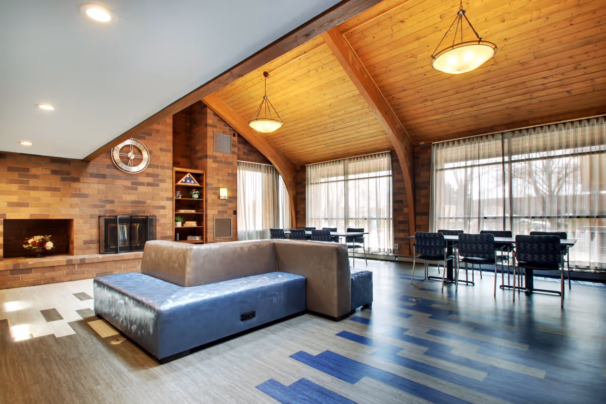 A spacious common area with a high wooden ceiling and large windows covered with sheer curtains. The room features a modern L-shaped sofa with blue and beige upholstery, a brick wall with a fireplace, a clock, and built-in shelves. Several tables and chairs are arranged near the windows, creating a comfortable and inviting lounge space.