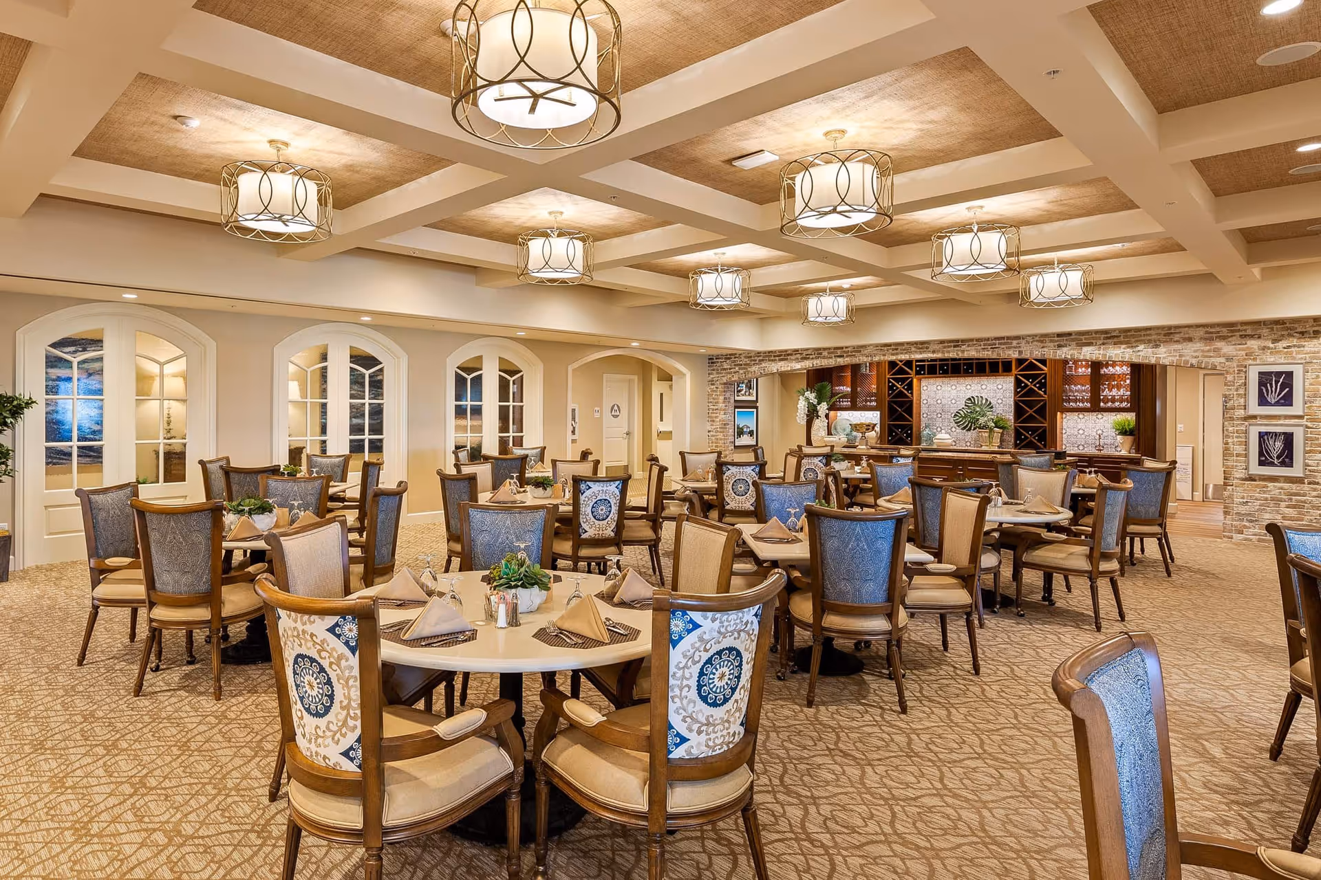 A spacious dining room with multiple round tables set with beige napkins and glassware. The chairs have wooden frames with patterned upholstery in blue and beige tones. The ceiling features recessed lighting and decorative hanging light fixtures. The walls are light-colored with arched windows and a brick accent wall behind a wooden cabinet and counter area, decorated with plants and framed artwork.
