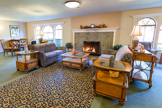 Bright, cozy common living room with upholstered sofas, wooden side tables, a lit fireplace, and a patterned area rug.