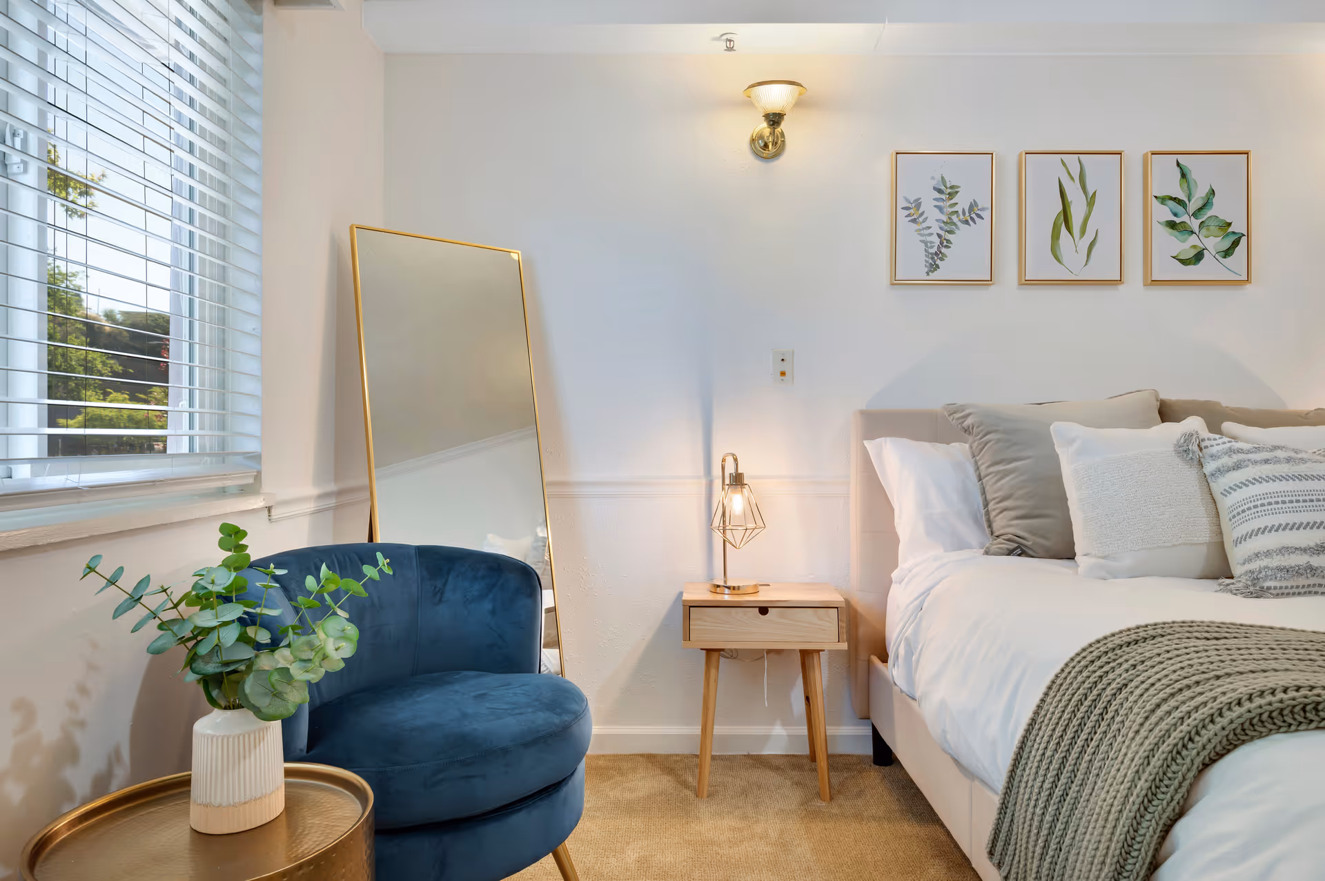 A cozy bedroom corner featuring a beige upholstered bed with multiple pillows and a knitted throw blanket. Next to the bed is a small wooden nightstand with a modern lamp. A large floor mirror with a gold frame leans against the wall. A blue velvet chair sits beside a round gold side table holding a vase with green foliage. Three framed botanical prints hang on the white wall above the bed.