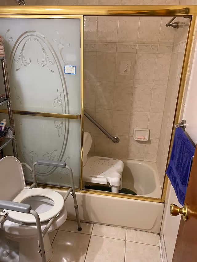 Bathroom with a toilet equipped with safety rails and a bathtub with a sliding glass door. Inside the bathtub is a white shower chair and a grab bar on the tiled wall. A blue towel hangs on a hook on the door.