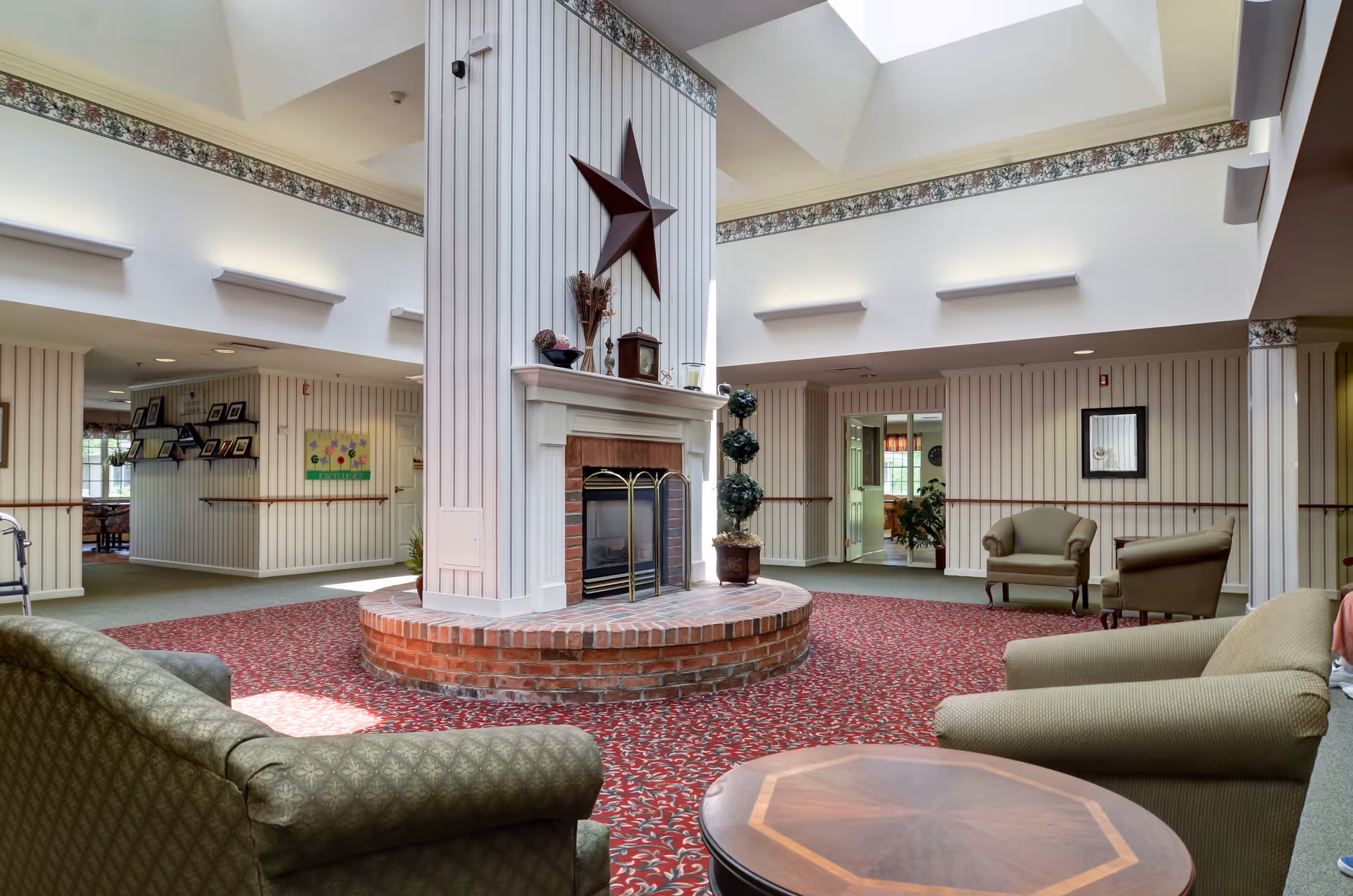 A spacious senior living facility common area with a central brick fireplace surrounded by a white wooden column featuring a large decorative star. The room has patterned red carpet, beige armchairs, and a wooden coffee table. The walls are decorated with vertical striped wallpaper and floral border trim near the ceiling. Skylights provide natural light, and there are doorways leading to other rooms.