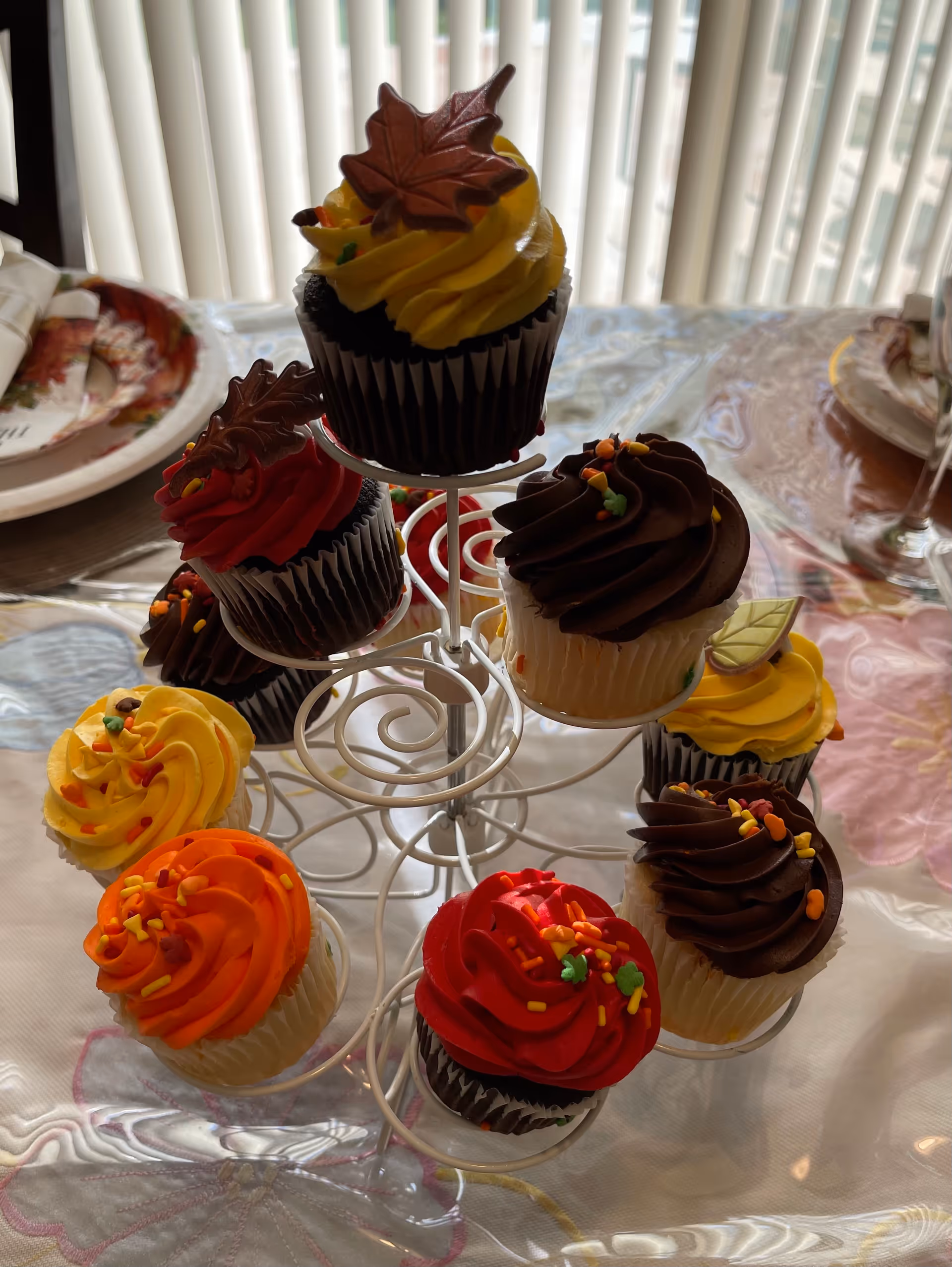 A white metal cupcake stand holding nine cupcakes decorated with colorful autumn-themed frosting in shades of yellow, orange, red, and chocolate brown, topped with leaf-shaped decorations. The stand is placed on a table with a floral tablecloth and plates in the background near vertical blinds.