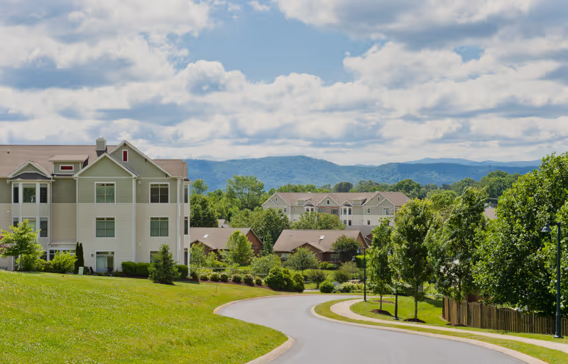 A winding road leading through a residential area with multiple buildings surrounded by green trees and grass, with mountains visible in the background under a partly cloudy sky.
