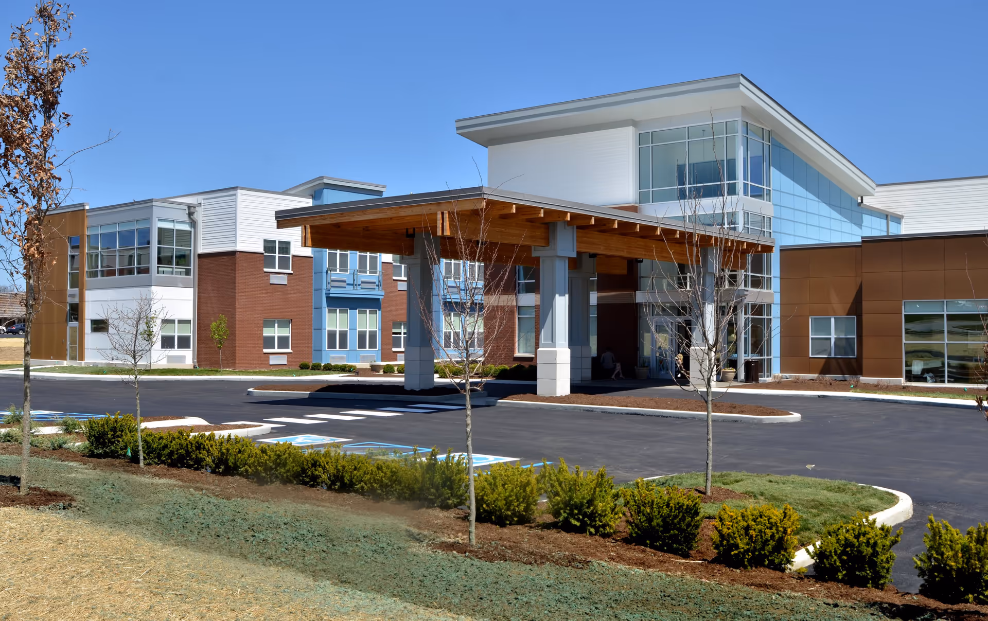 Exterior view of a modern senior living facility building with a covered entrance, large windows, and a landscaped area with small trees and bushes under a clear blue sky.