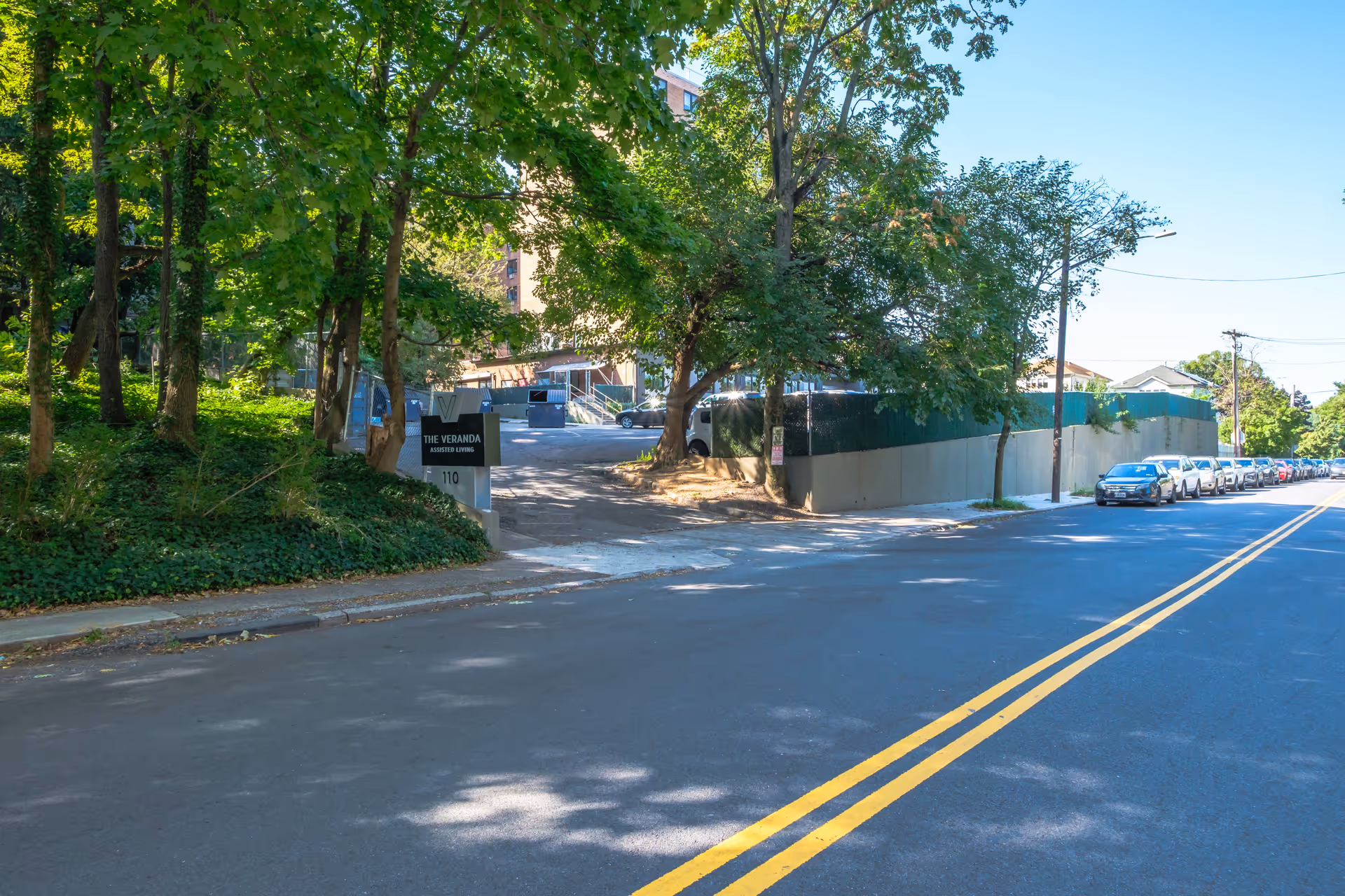 Street view showing the entrance and sign for The Veranda Assisted Living, a tree-lined driveway and parked cars along the road.
