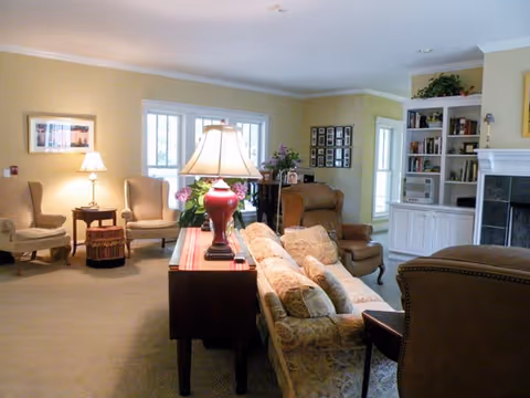 A cozy living room with beige walls and carpet, featuring a floral patterned sofa, several armchairs, a wooden side table with a lamp, built-in white shelves with books and decorations, and a fireplace with a black tile surround.