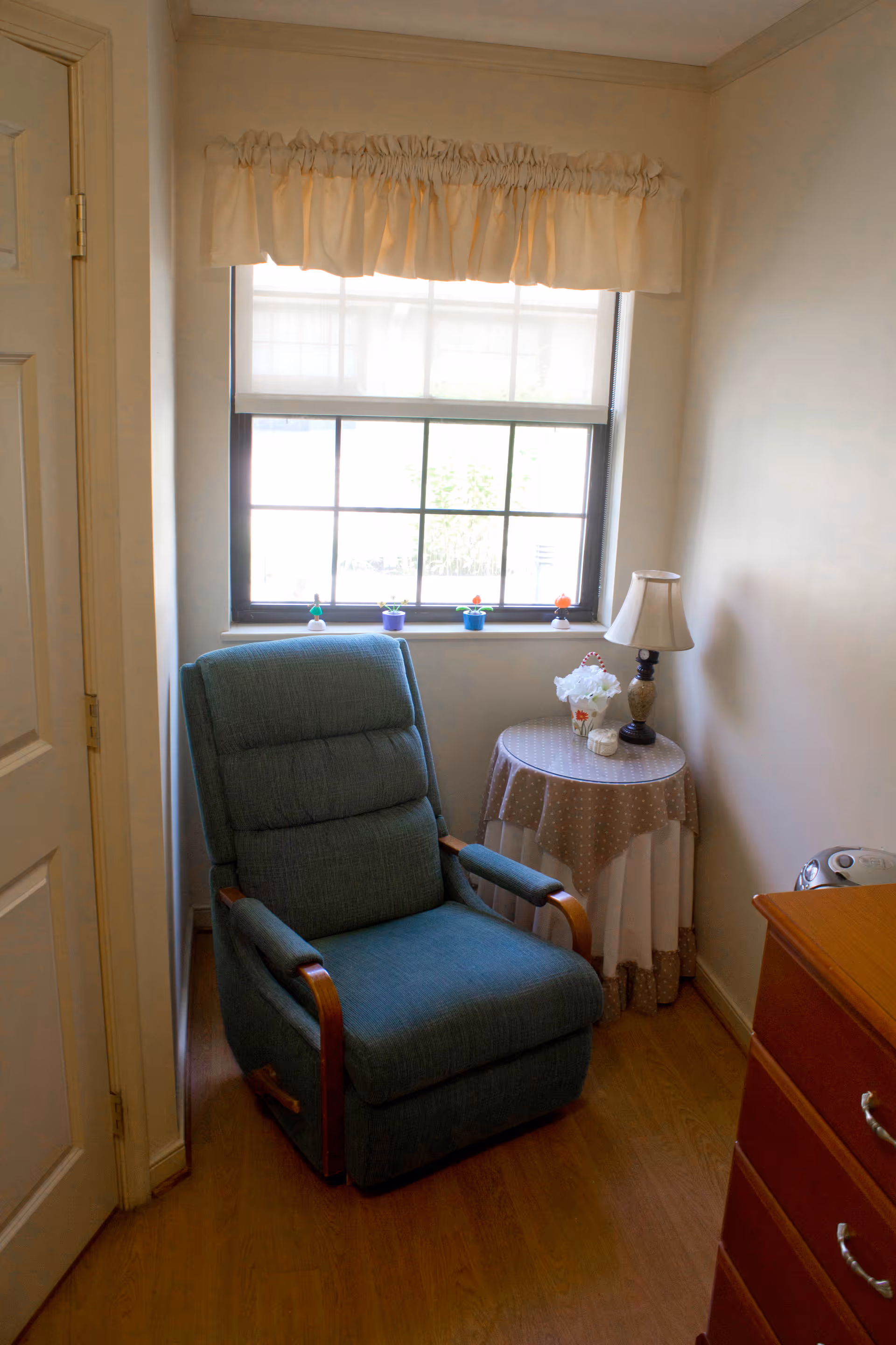 A cozy corner of a room featuring a blue upholstered recliner chair positioned near a window with a white valance and a roller shade. Next to the chair is a round table covered with a polka dot tablecloth, holding a small lamp and decorative items. The floor is wooden, and part of a wooden dresser is visible on the right side.