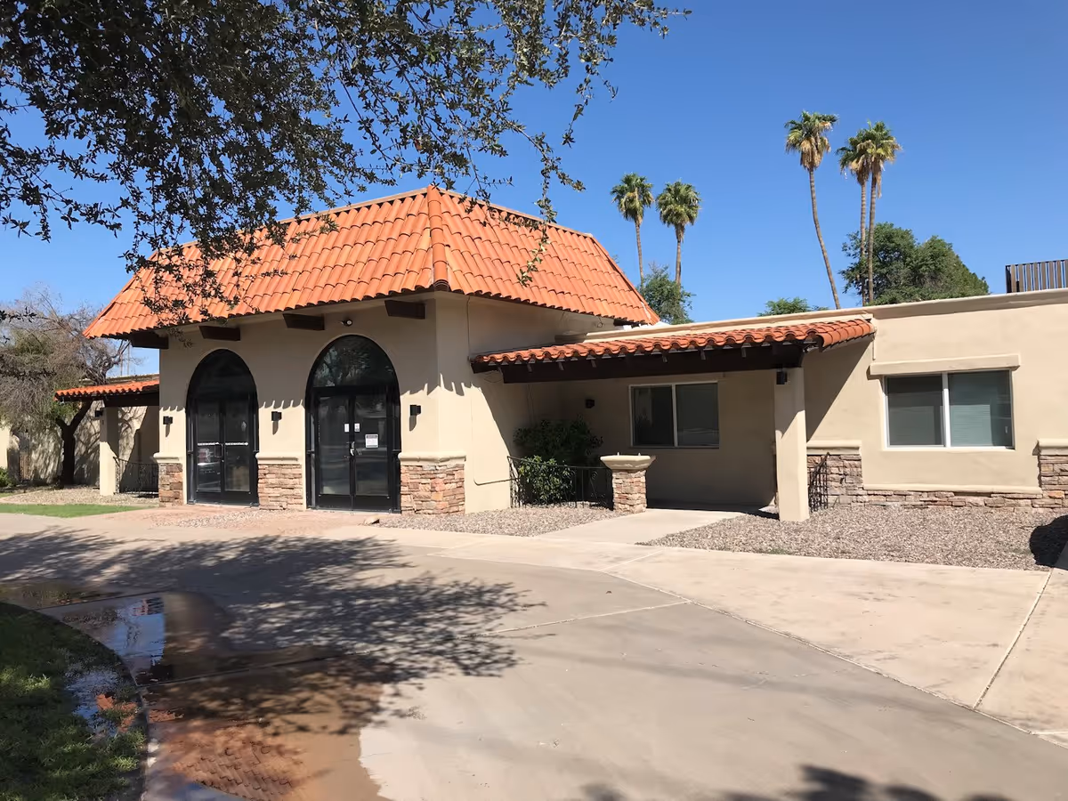 Front entrance of a one-story beige building with a red tile roof, arched glass doors, and palm trees in the background.