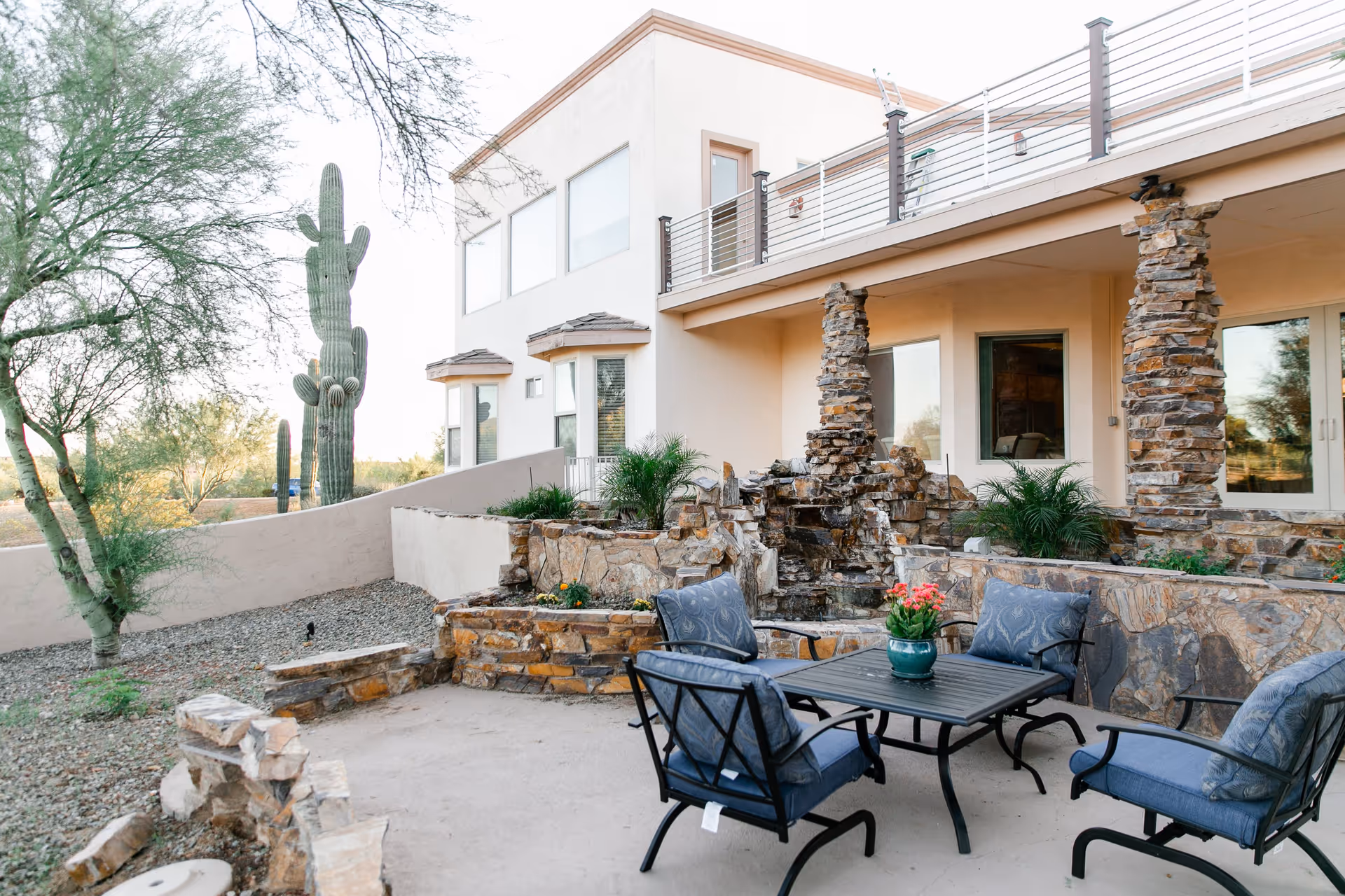 Stone patio with a table and four cushioned chairs in front of a two-story stucco building with stone columns, desert landscaping and a large saguaro cactus.