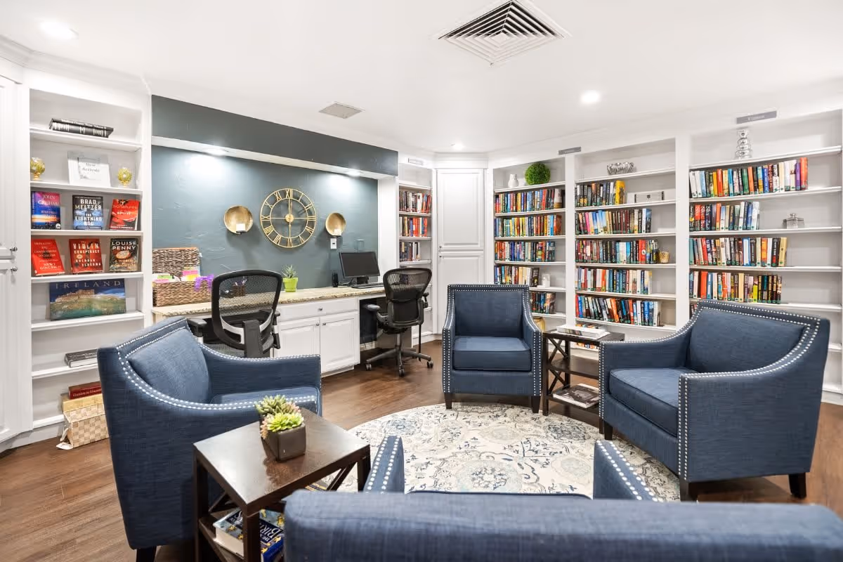 A cozy library room with white built-in bookshelves filled with books, four blue upholstered armchairs arranged around a circular patterned rug, a small wooden side table with a plant, and a desk area with two office chairs, a computer, and a decorative wall clock.