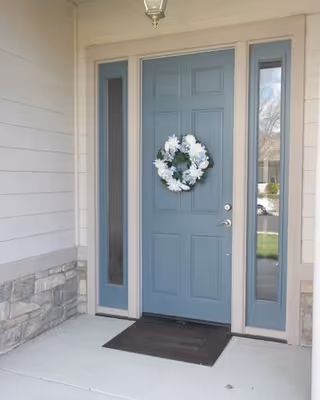Blue paneled front door with a white floral wreath, flanked by narrow glass sidelights on a covered porch with stone trim and a black doormat.