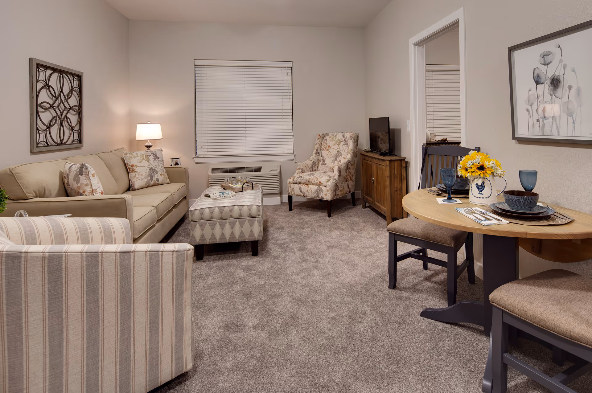 A cozy living room with a beige sofa, a striped armchair, a patterned armchair, and a cushioned ottoman. There is a wooden TV stand with a flat-screen TV, a small dining table set for two with blue glasses and a vase of yellow flowers, and wall decorations including a metal art piece and a framed floral painting. The room has carpeted flooring and a window with closed blinds.