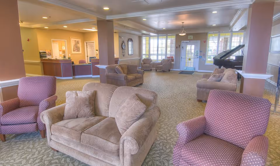 A spacious assisted living facility common area with multiple upholstered armchairs and sofas arranged on a patterned carpet. The room features large windows allowing natural light, a grand piano in the corner, and a reception desk in the background.