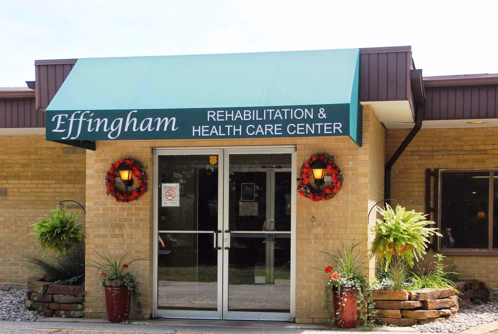 Entrance to Effingham Rehabilitation & Health Care Center with double glass doors under a green awning. Two red wreaths with lights are mounted on the brick wall on either side of the doors. There are potted plants and hanging ferns decorating the entrance area.