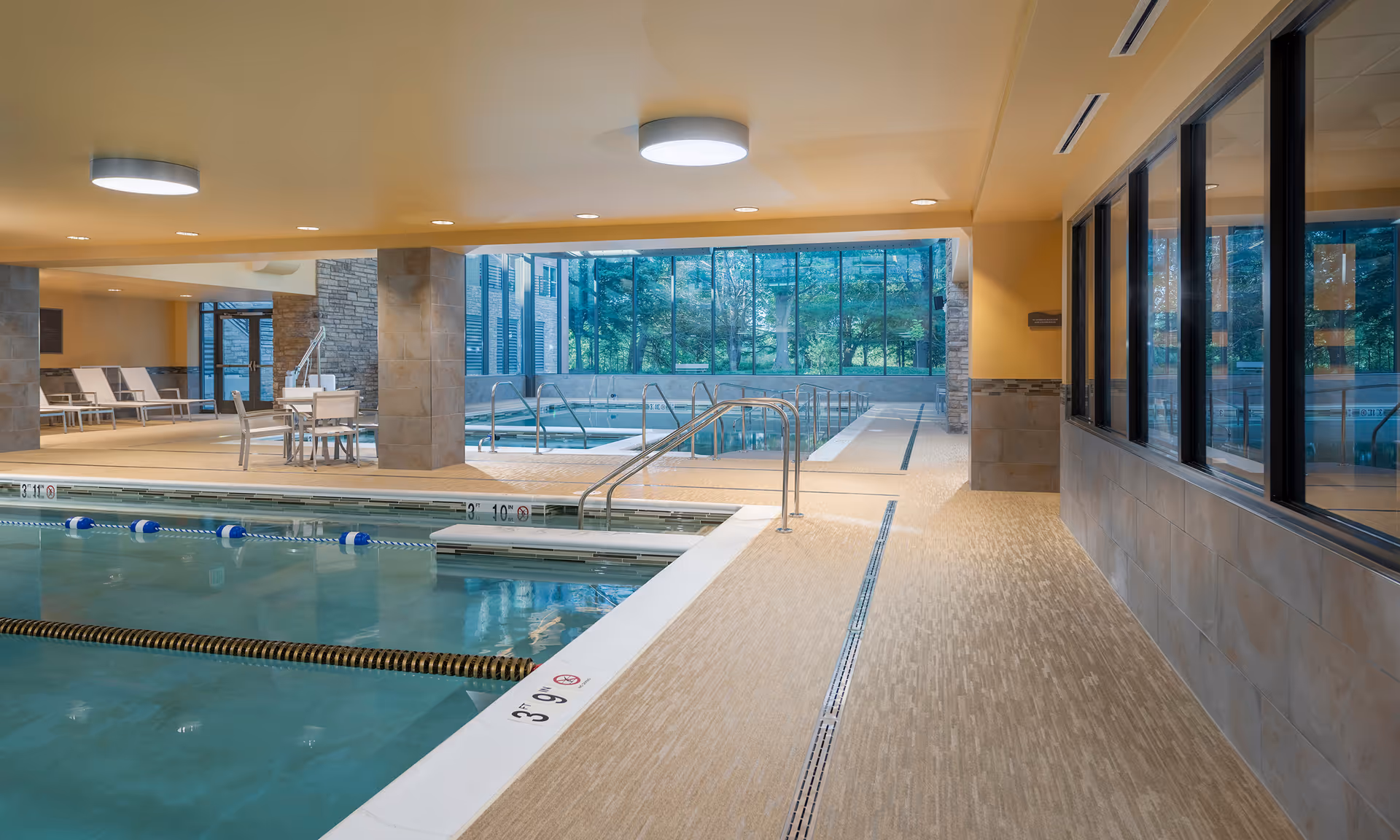 Indoor swimming pool area with clear water, lane dividers, and depth markings. The pool is surrounded by beige tiled flooring and walls with large windows providing a view of greenery outside. There are lounge chairs and a table with chairs near the pool, and ceiling lights illuminate the space.