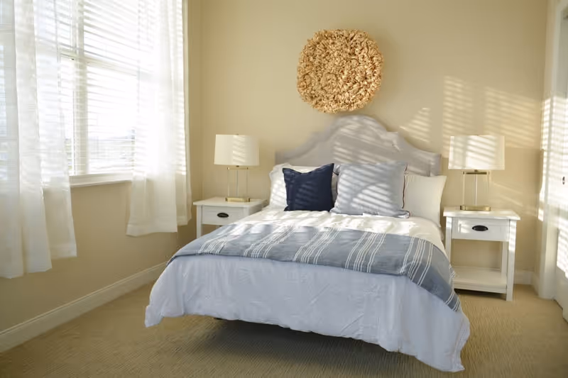A bright bedroom with a neatly made bed featuring white and blue bedding, two white nightstands with matching lamps, and a decorative wall hanging above the headboard. Sunlight streams through sheer white curtains on a window to the left.