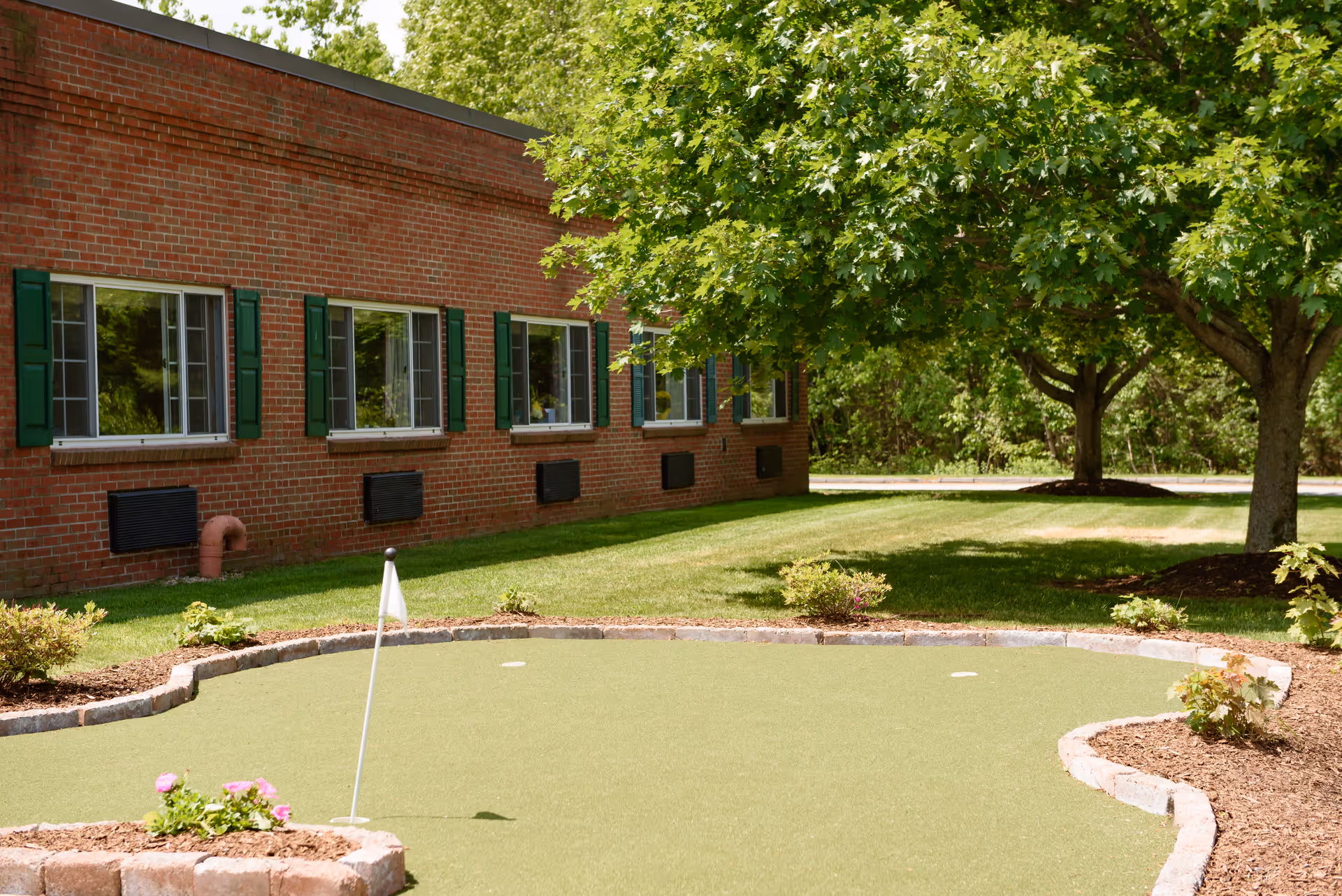 A small putting green and landscaped lawn beside a brick senior living building with green-shuttered windows and trees.