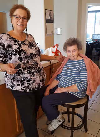 An elderly woman with a pink towel draped over her shoulders sits on a stool in a kitchen area, smiling. A younger woman with glasses and curly hair stands next to her, also smiling, holding a comb. The setting appears to be a home or assisted living facility kitchen with cabinets and a calendar on the wall.