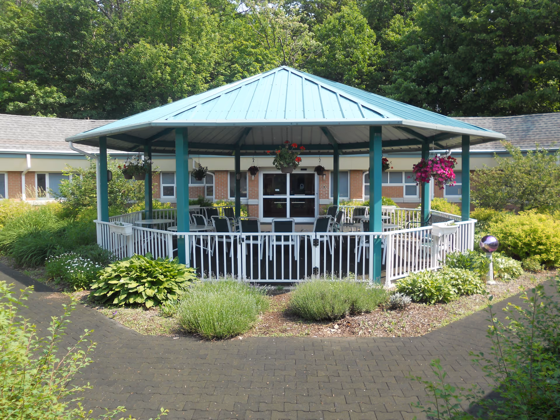 A gazebo with a teal metal roof and green support beams surrounded by a white railing. Inside the gazebo are several black chairs and tables. Hanging flower baskets with red and purple flowers are attached to the gazebo. The gazebo is situated in a garden area with various green shrubs and plants, and a paved pathway leads up to it. In the background, there is a single-story brick building with windows and a door.