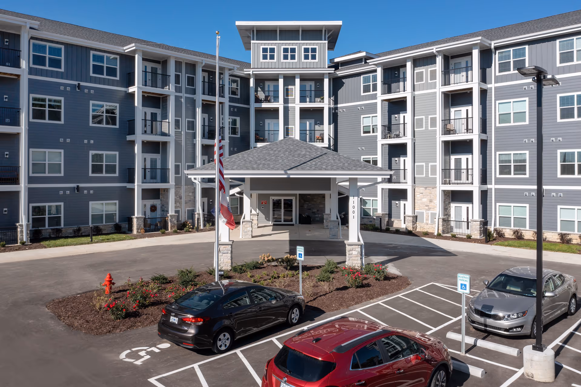 Exterior view of Avalon Park Independent Living facility showing a modern multi-story building with balconies, a covered entrance, an American flag, landscaped flower beds, and a parking lot with several cars including handicapped parking spaces.