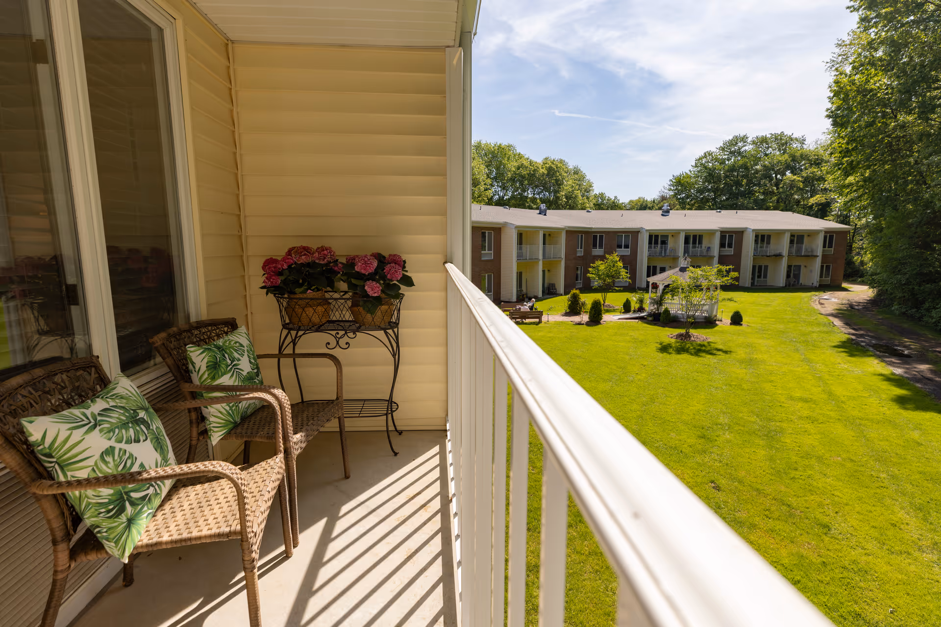 A sunny balcony with two wicker chairs featuring green leaf-patterned cushions and a small table holding a basket of pink flowers. The balcony overlooks a well-maintained grassy courtyard with trees, shrubs, benches, and a gazebo. In the background, there is a two-story building with balconies and windows.